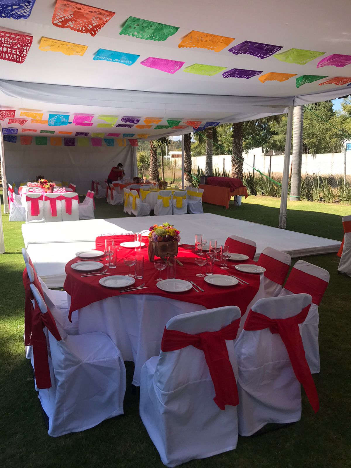 Festejo con papel picado multicolor bajo carpa, mesas en rojo y amarillo con pista de baile para XV años o boda